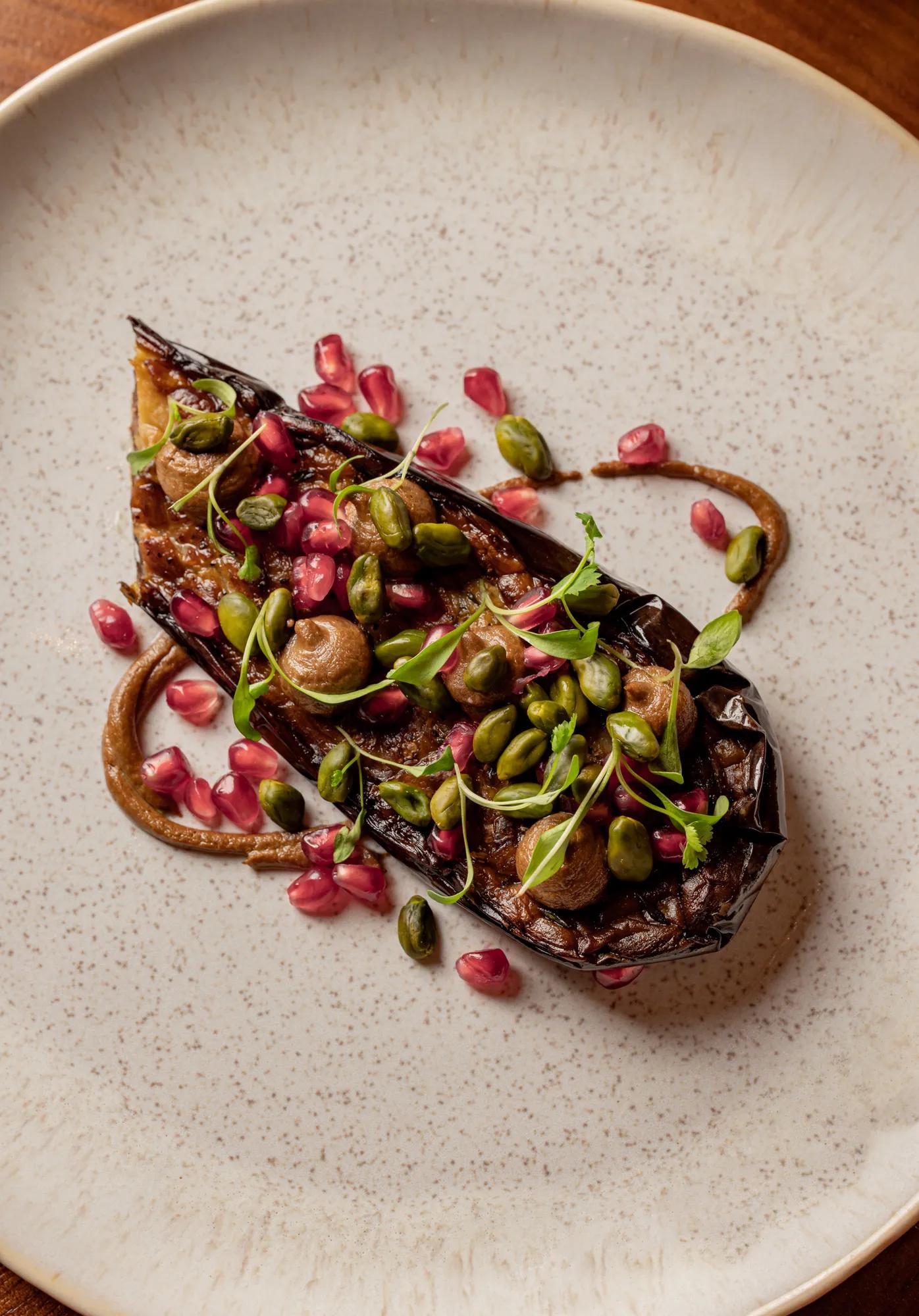 Close-up of a beautifully plated dish featuring roasted eggplant topped with pomegranate seeds, green pistachios, and fresh herbs, arranged artfully on a textured plate.