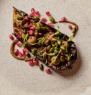 Close-up of a beautifully plated dish featuring roasted eggplant topped with pomegranate seeds, green pistachios, and fresh herbs, arranged artfully on a textured plate.