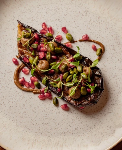 Close-up of a beautifully plated dish featuring roasted eggplant topped with pomegranate seeds, green pistachios, and fresh herbs, arranged artfully on a textured plate.