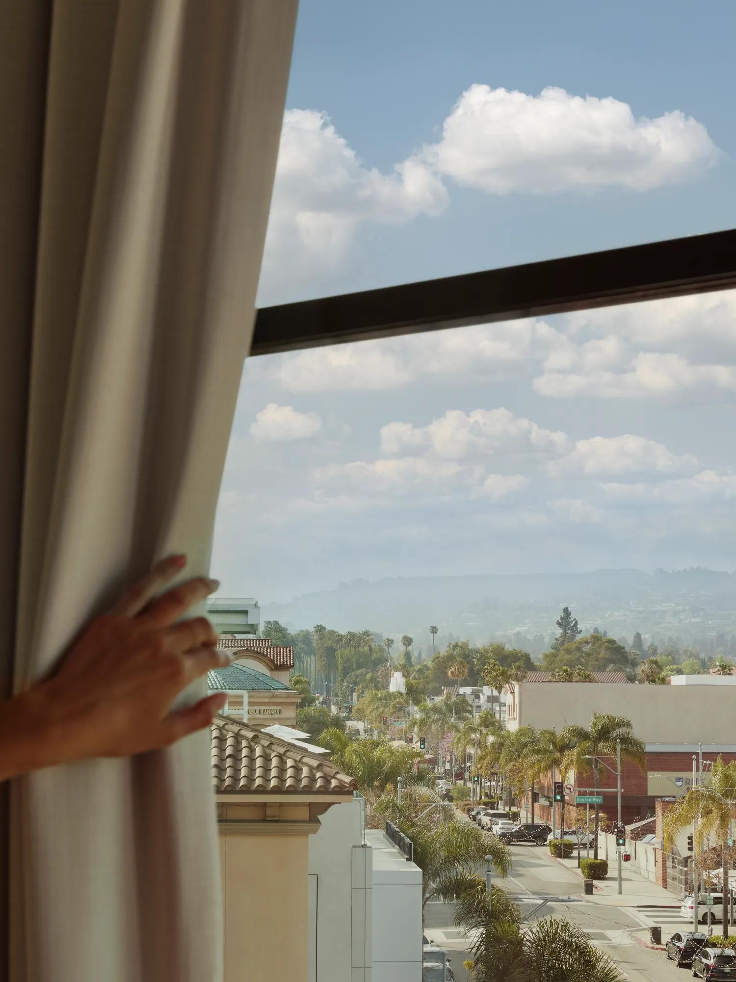 View through a hotel room window overlooking a palm-lined city street and distant hills, framed by curtains under a bright, cloudy sky.