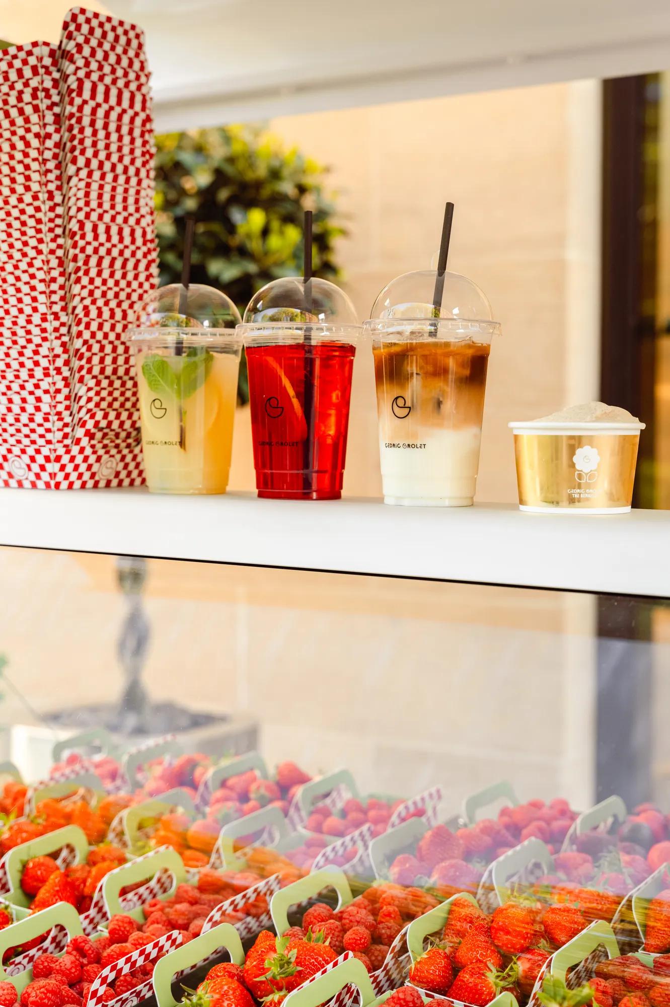 Display of takeaway beverages including iced citrus drinks, iced coffee and a dessert cup, arranged above baskets of fresh strawberries at a market-style counter.