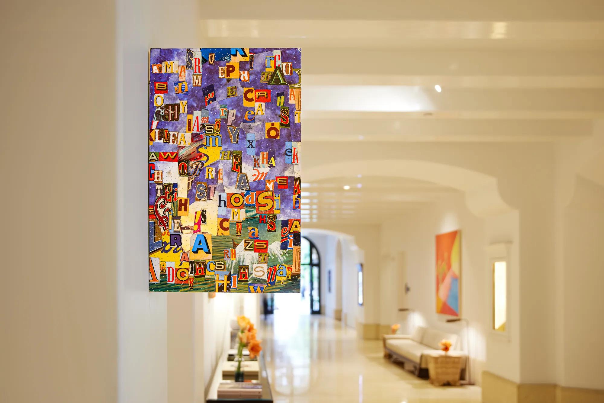 Colorful abstract artwork made from scattered letters and typographic cutouts mounted on a wall in a bright, modern hallway. The artwork features overlapping letters in various sizes, colors, and fonts against a predominantly purple background. In the softly blurred corridor behind it are white arches, polished floors, a bench, and additional abstract artwork displayed along the walls.