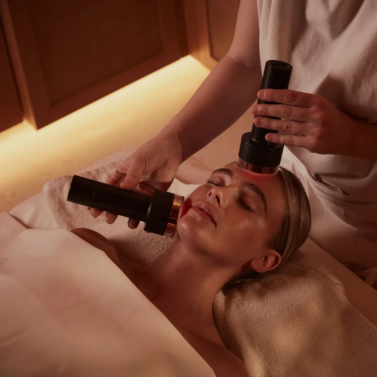 Person receiving a facial treatment at a spa with handheld devices applied to their face, while lying on a treatment bed covered in a towel, with soft lighting in the background.