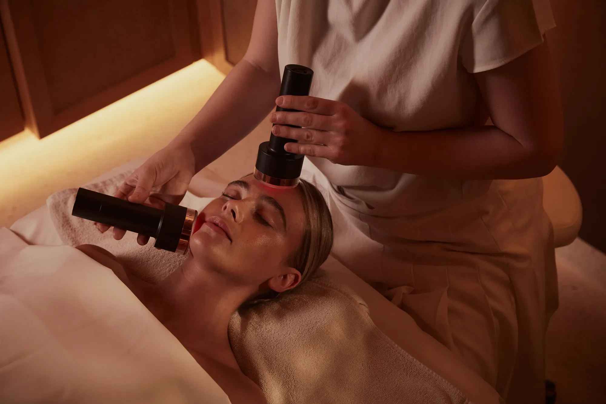 Person receiving a facial treatment at a spa with handheld devices applied to their face, while lying on a treatment bed covered in a towel, with soft lighting in the background.