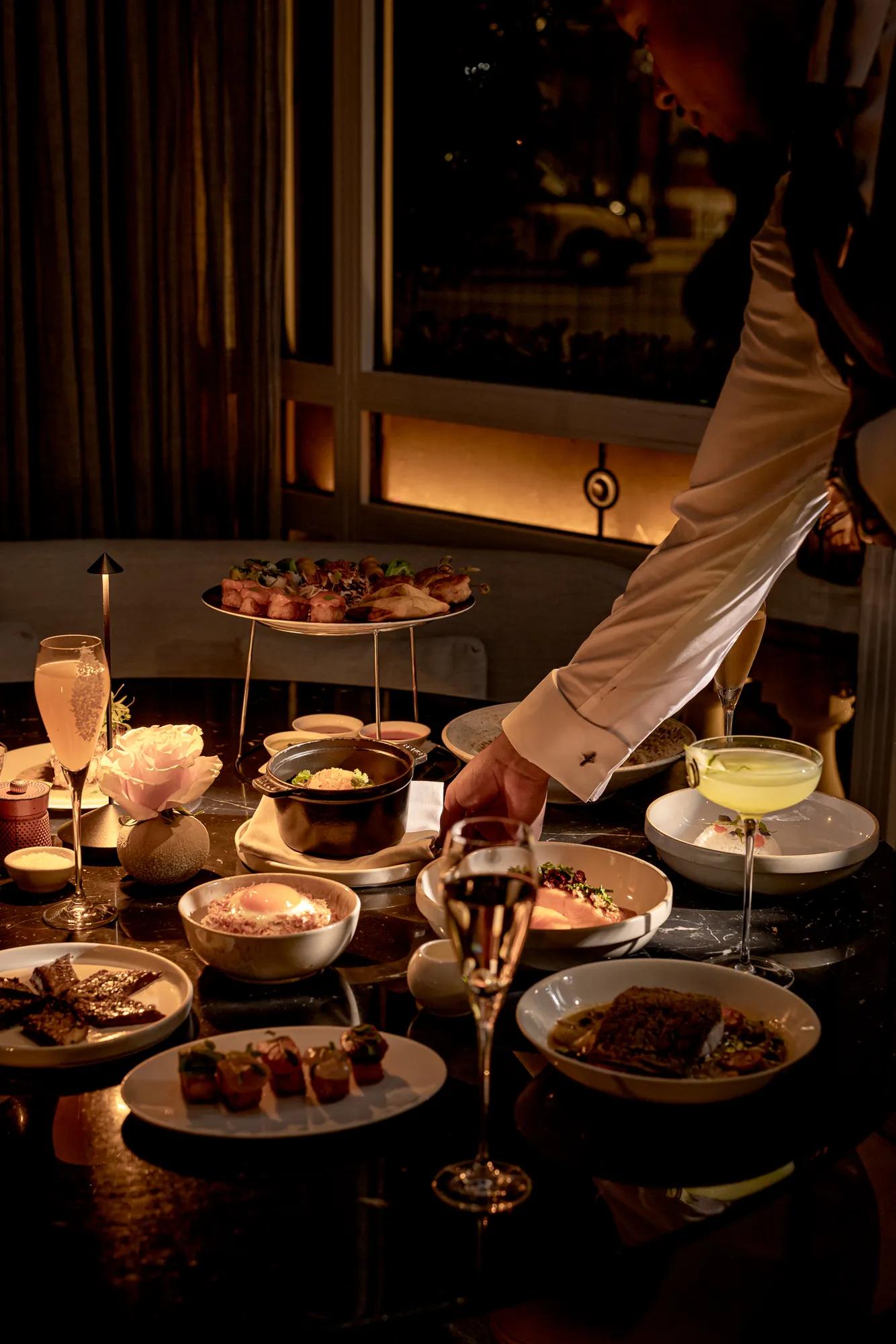 Server placing dishes on a table set with cocktails and a selection of sharing plates in a softly lit dining room.