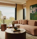 Living area with a textured beige sofa, round marble-topped coffee table, modern side tables, and large windows overlooking trees, styled with warm wood panelling and artwork.
