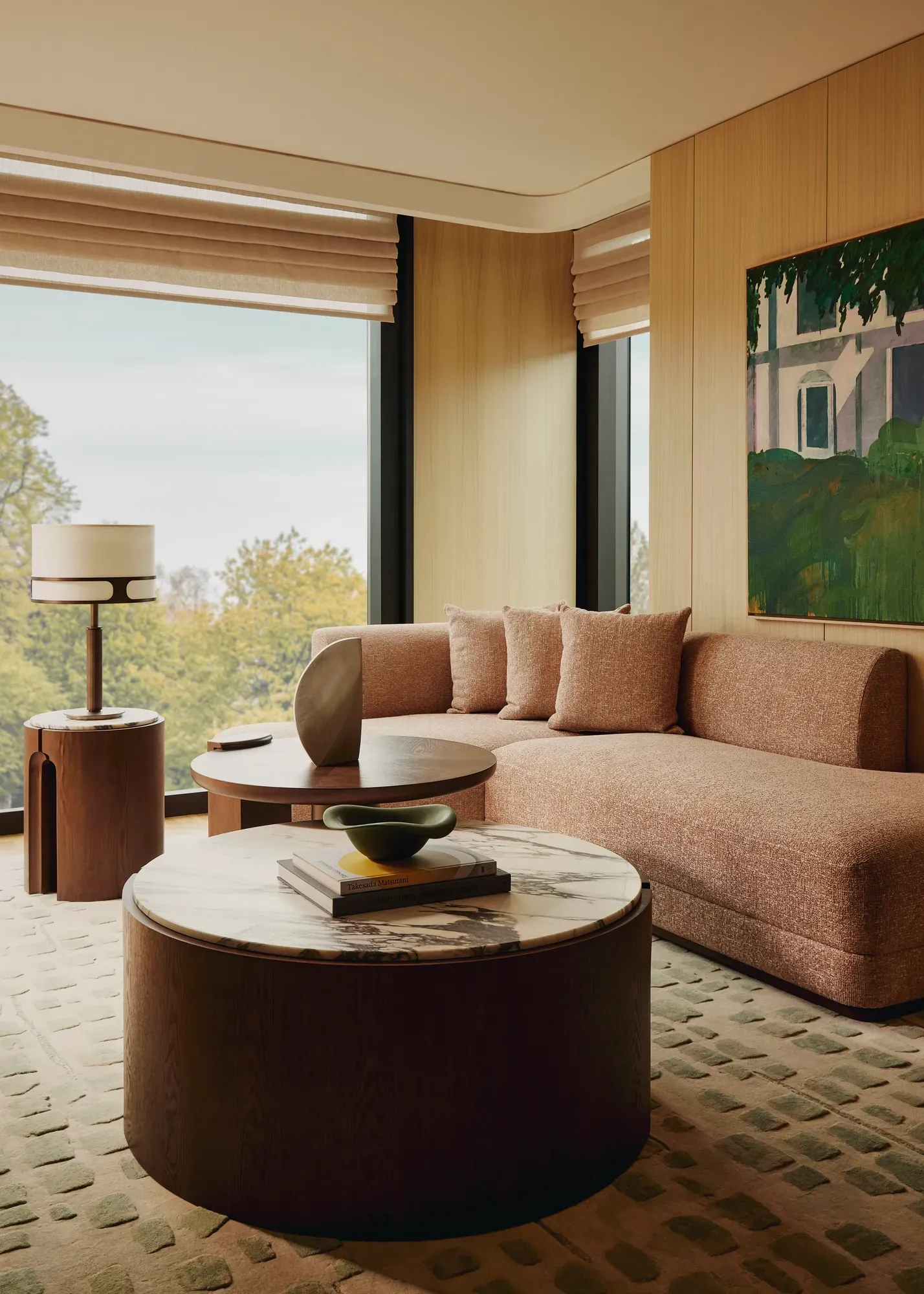 Living area with a textured beige sofa, round marble-topped coffee table, modern side tables, and large windows overlooking trees, styled with warm wood panelling and artwork.