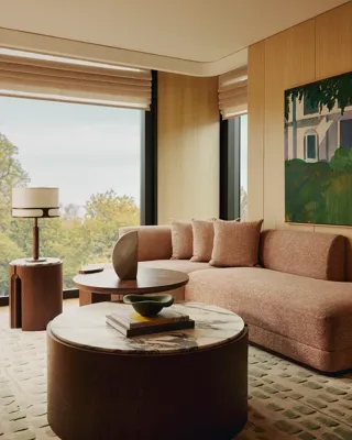 Living area with a textured beige sofa, round marble-topped coffee table, modern side tables, and large windows overlooking trees, styled with warm wood panelling and artwork.