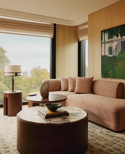 Living area with a textured beige sofa, round marble-topped coffee table, modern side tables, and large windows overlooking trees, styled with warm wood panelling and artwork.