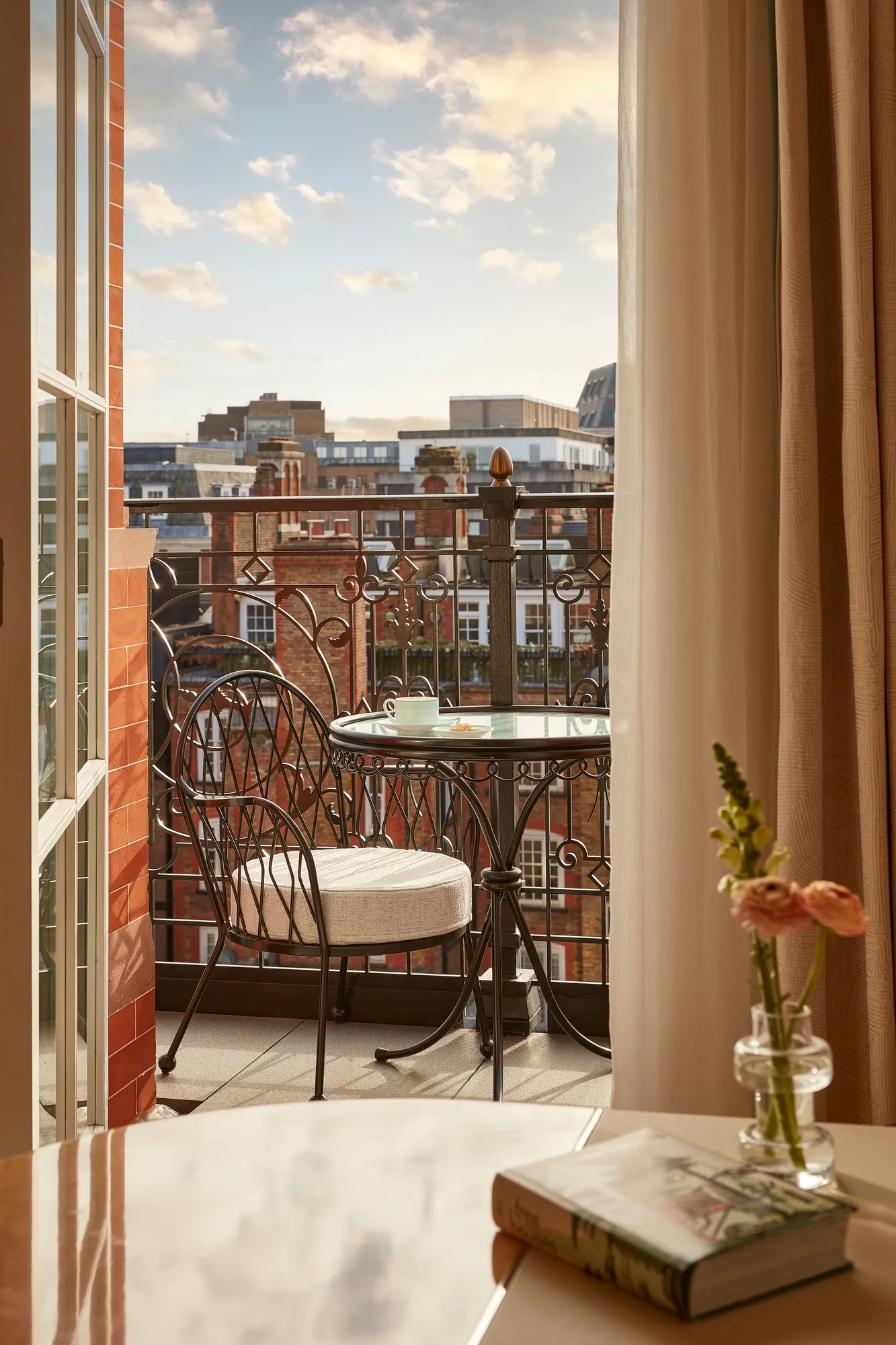 Balcony with wrought iron railing, a small round table, and two cushioned chairs overlooking red-brick buildings under a softly lit evening sky.