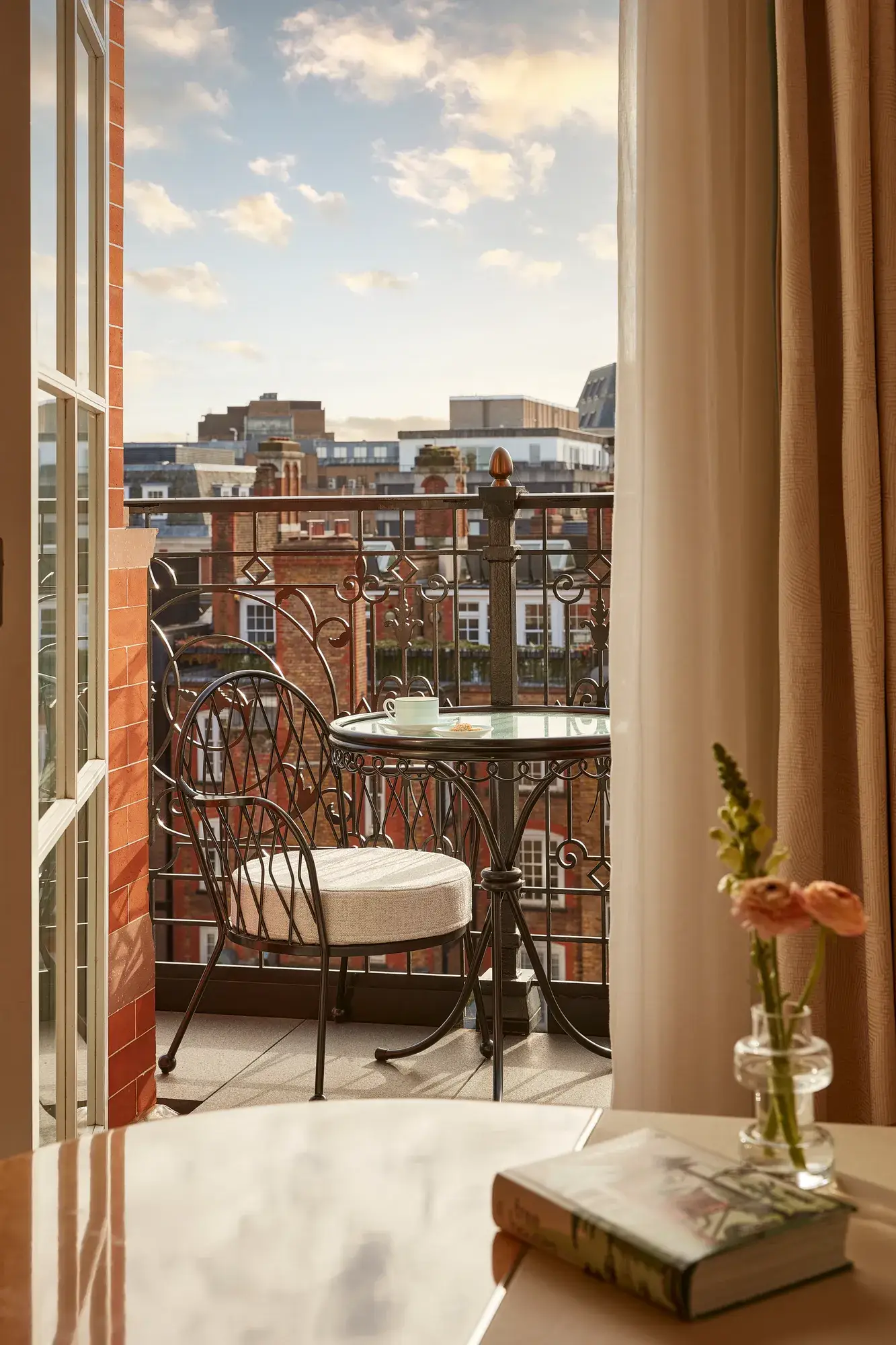Balcony with wrought iron railing, a small round table, and two cushioned chairs overlooking red-brick buildings under a softly lit evening sky.