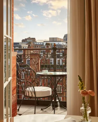 Balcony with wrought iron railing, a small round table, and two cushioned chairs overlooking red-brick buildings under a softly lit evening sky.