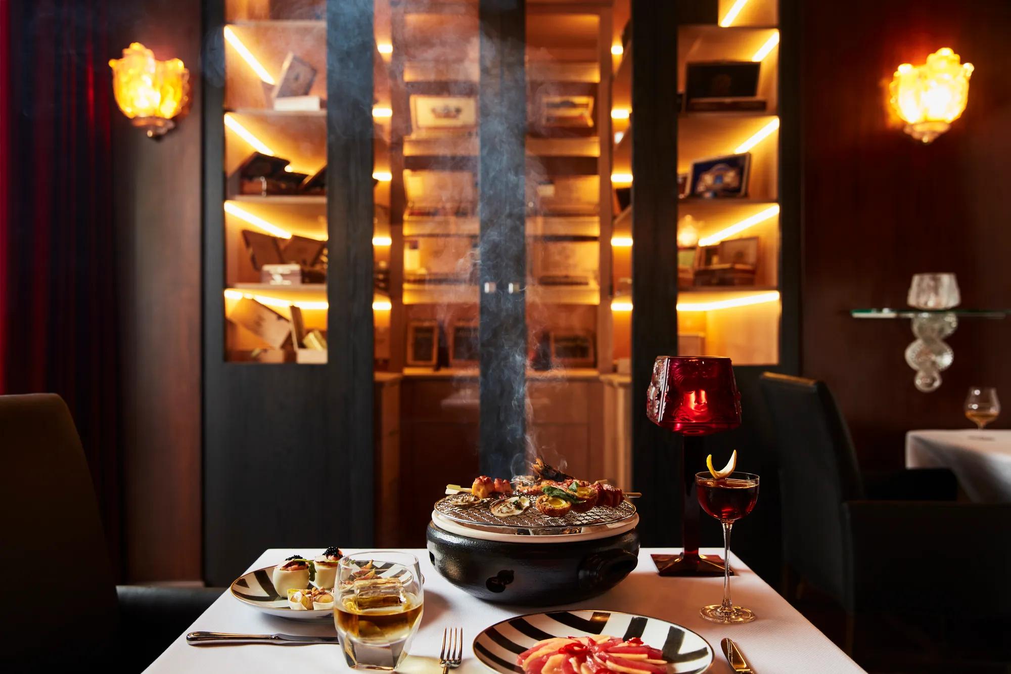 Table with tabletop grill, cocktails, and appetizers in dimly lit whisky bar.
