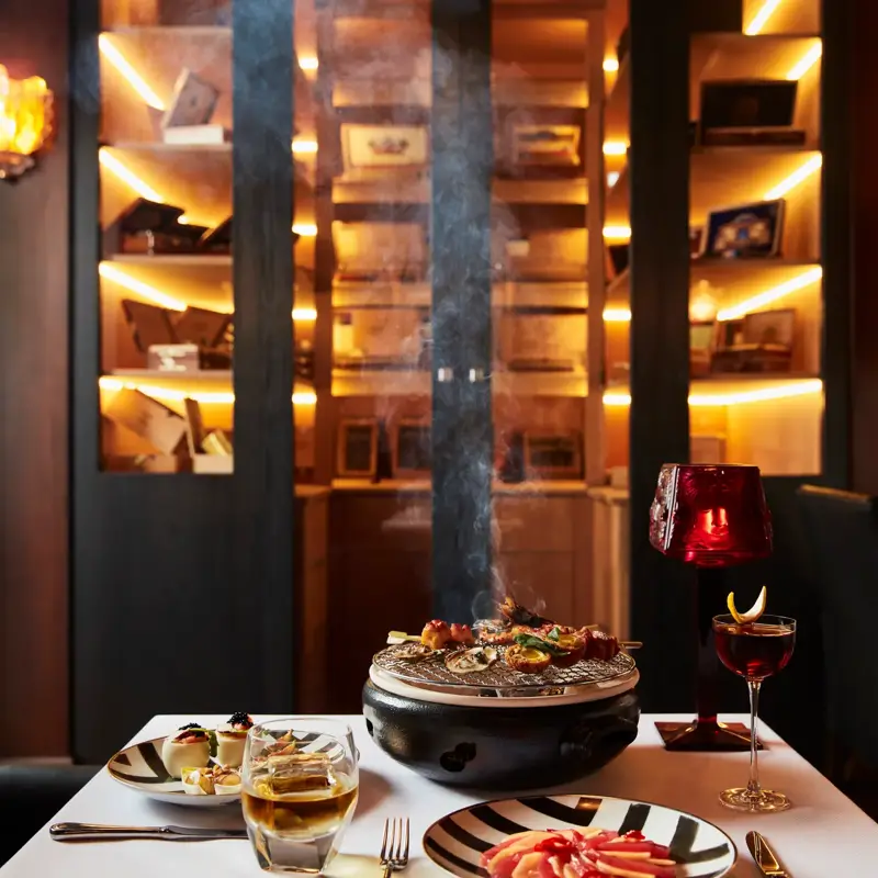 Table with tabletop grill, cocktails, and appetizers in dimly lit whisky bar.