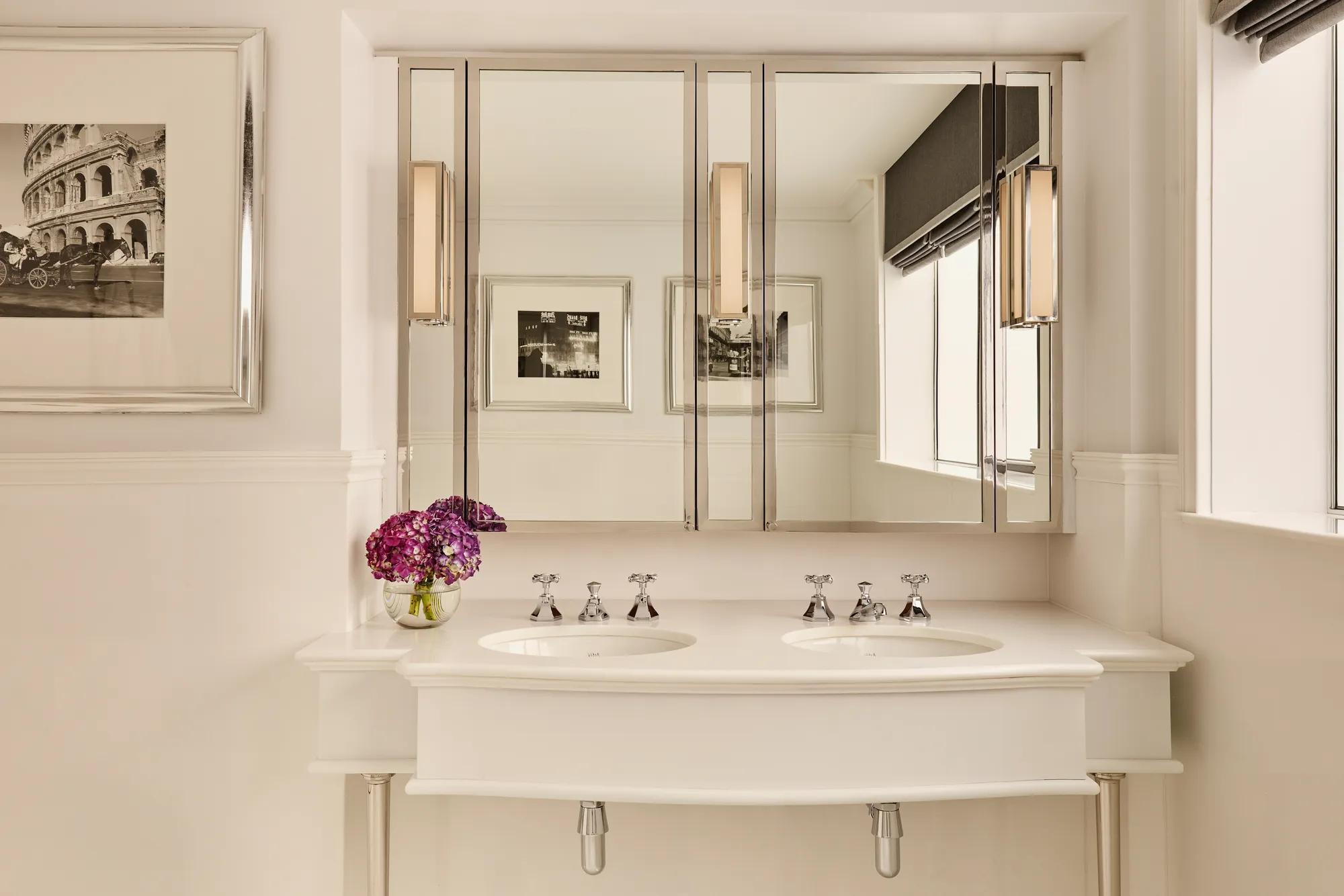 Cream double vanity with mirrored cabinets, chrome taps, and purple flowers beside bright window.