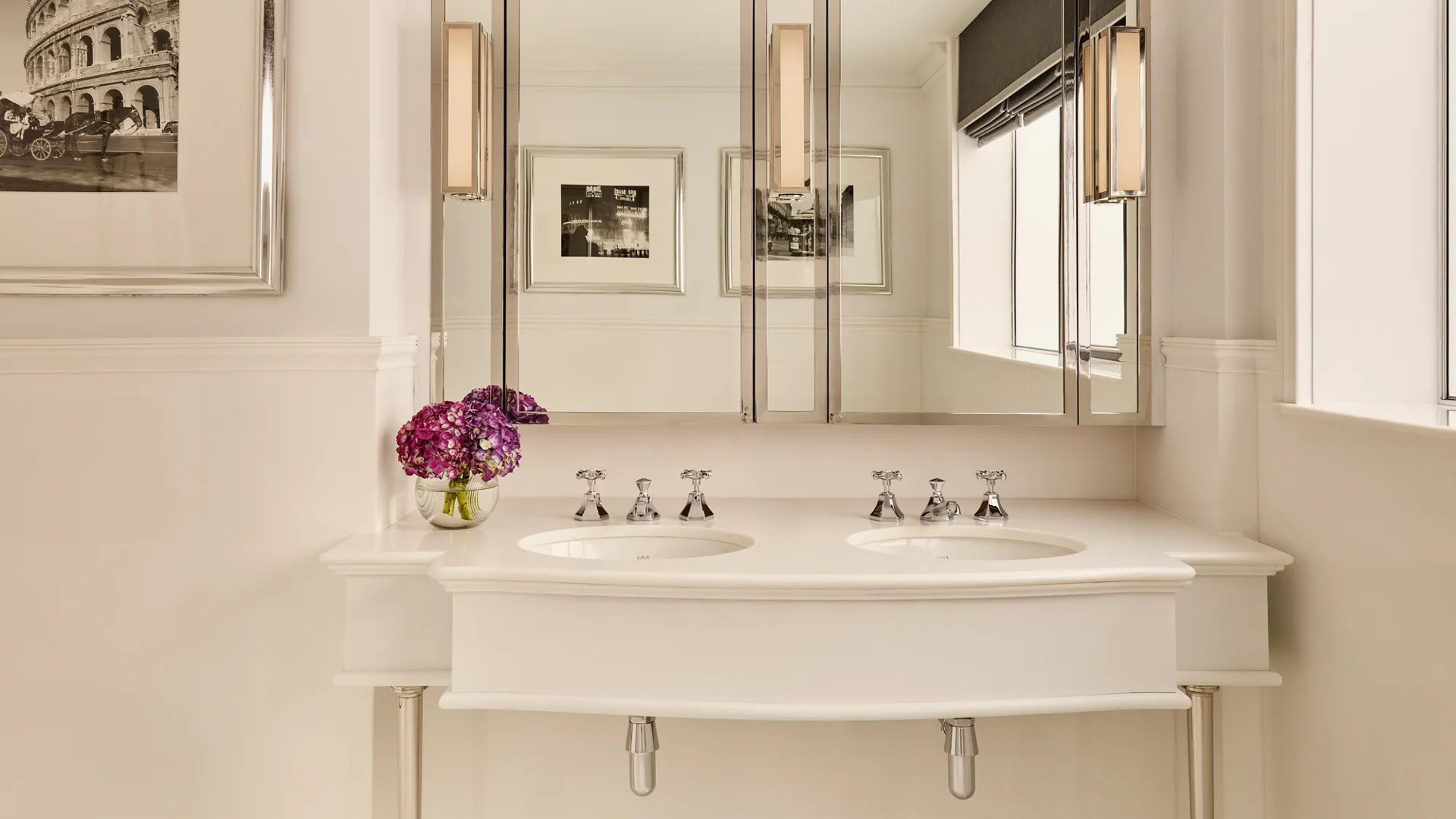 Cream double vanity with mirrored cabinets, chrome taps, and purple flowers beside bright window.