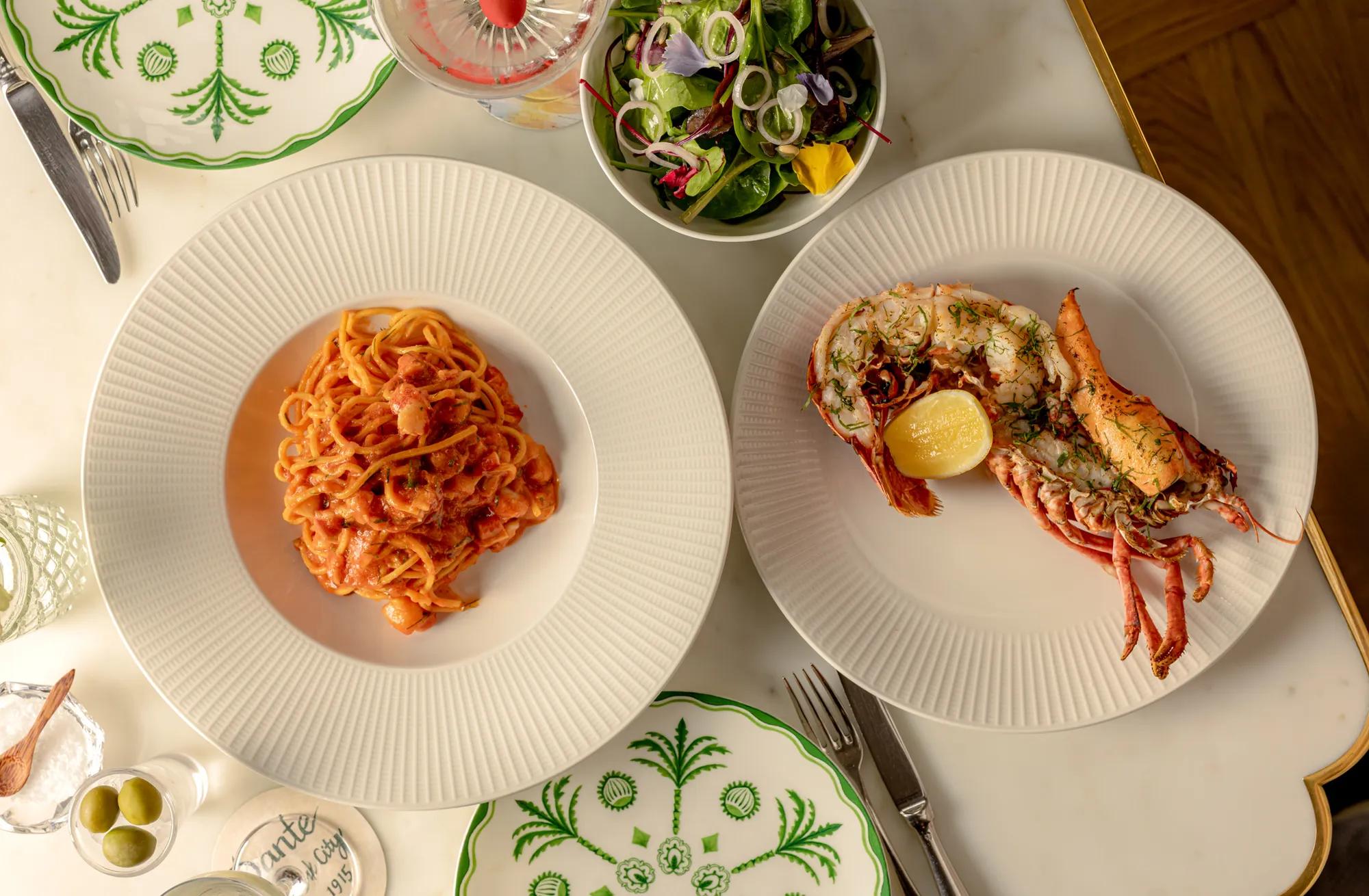 Lobster tail with lemon and herb garnish served alongside creamy tomato seafood pasta, with a mixed green salad on a marble dining table.