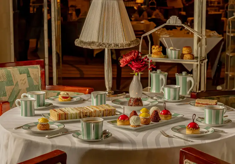 Afternoon tea table with pastries, sandwiches, striped teacups, and red floral centerpiece.
