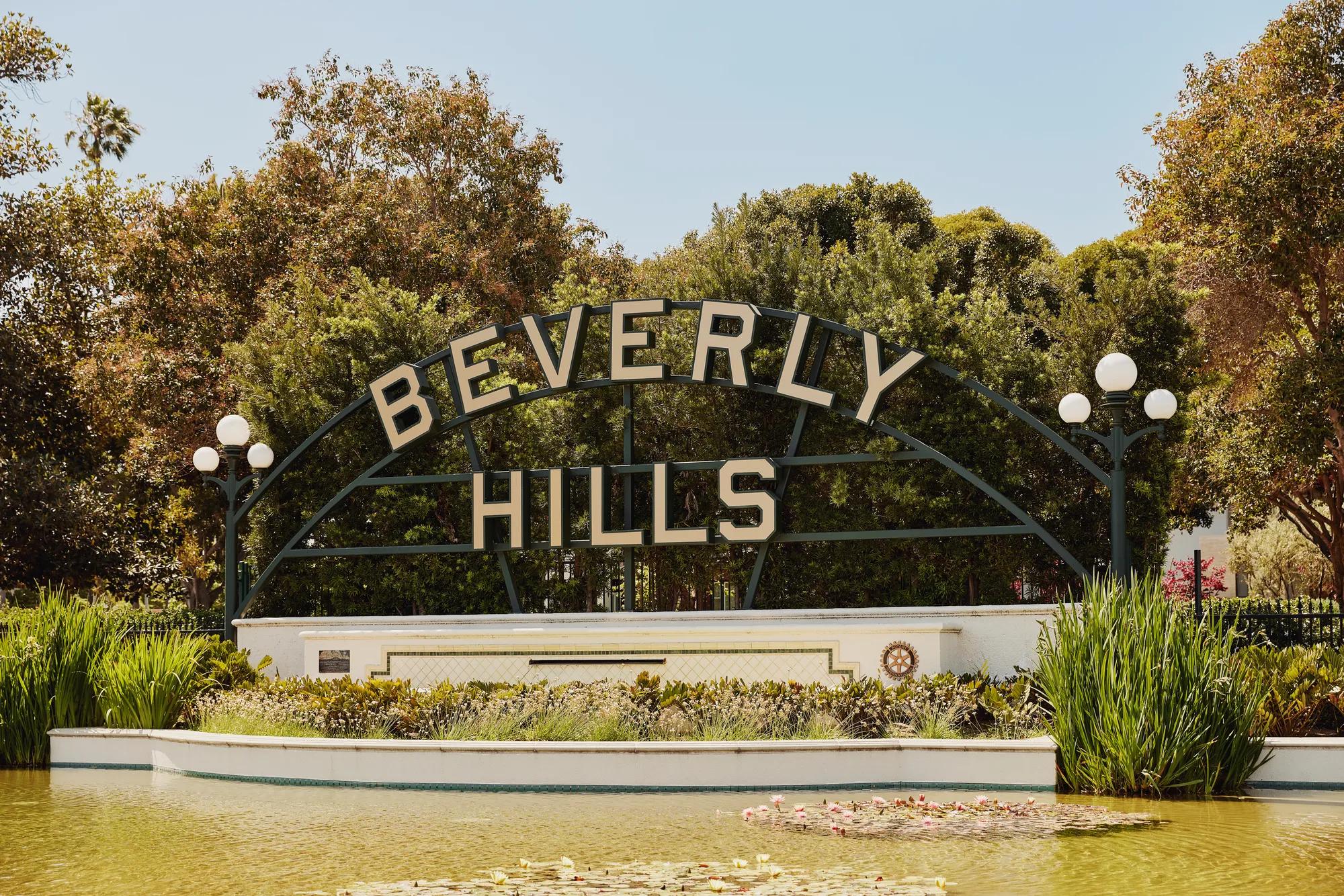 Beverly Hills park sign above a reflecting pond with lilies, globe lamps, and landscaped greenery on a sunny day.