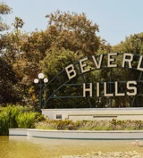 Beverly Hills park sign above a reflecting pond with lilies, globe lamps, and landscaped greenery on a sunny day.