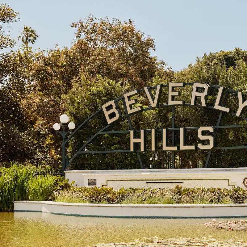 Beverly Hills park sign above a reflecting pond with lilies, globe lamps, and landscaped greenery on a sunny day.