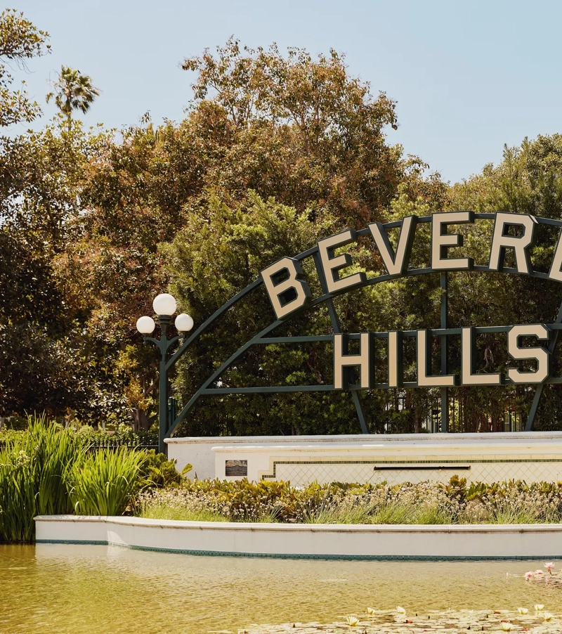 Beverly Hills park sign above a reflecting pond with lilies, globe lamps, and landscaped greenery on a sunny day.