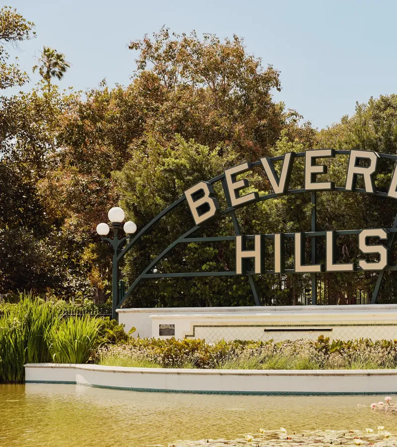 Beverly Hills park sign above a reflecting pond with lilies, globe lamps, and landscaped greenery on a sunny day.