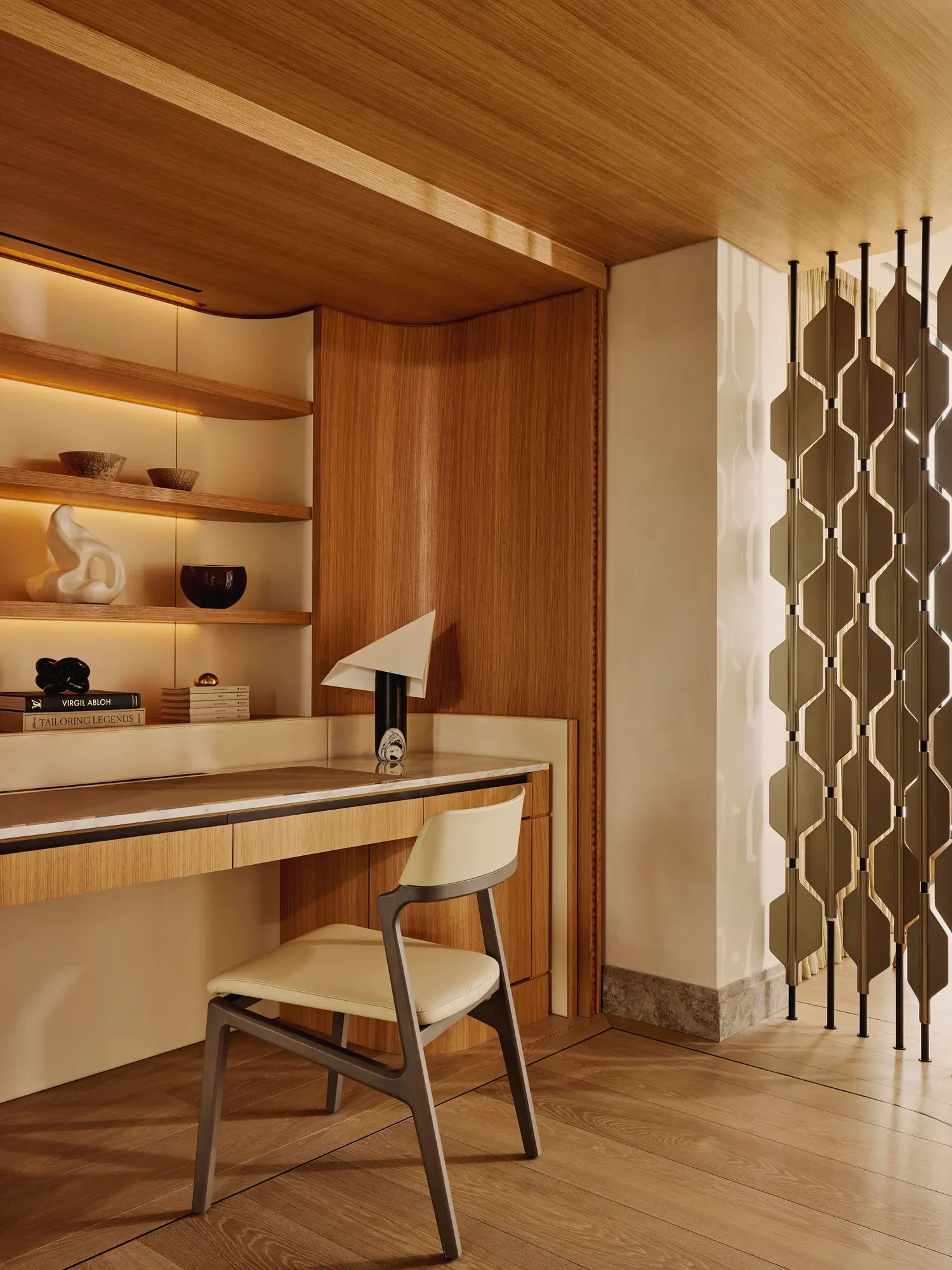 Minimalist writing desk with cream chair, open shelving and sculptural décor in a warm wood-panelled corner.
