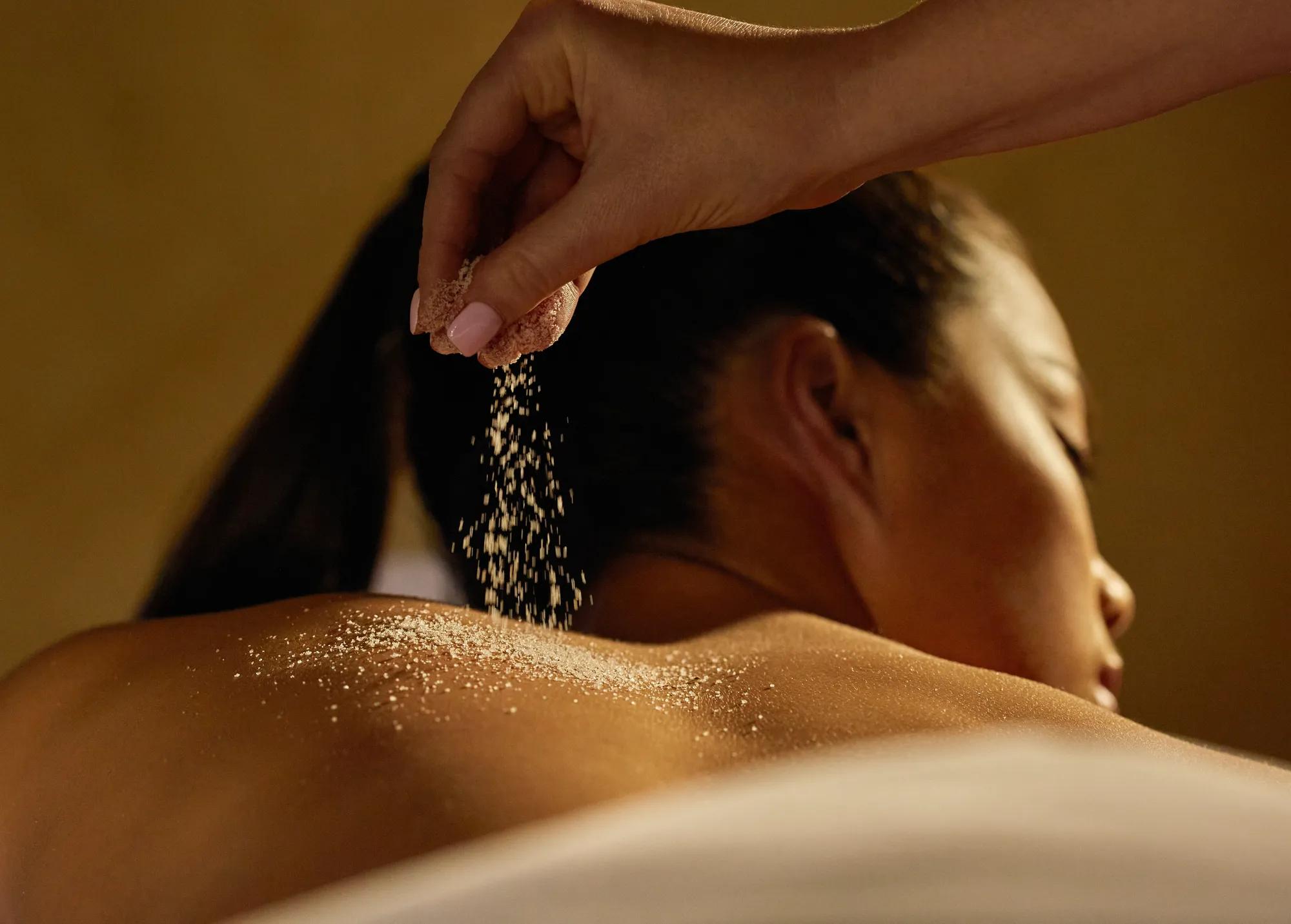 A hand sprinkles exfoliating salt onto a guest’s bare back during a spa treatment in a softly lit, relaxing setting.