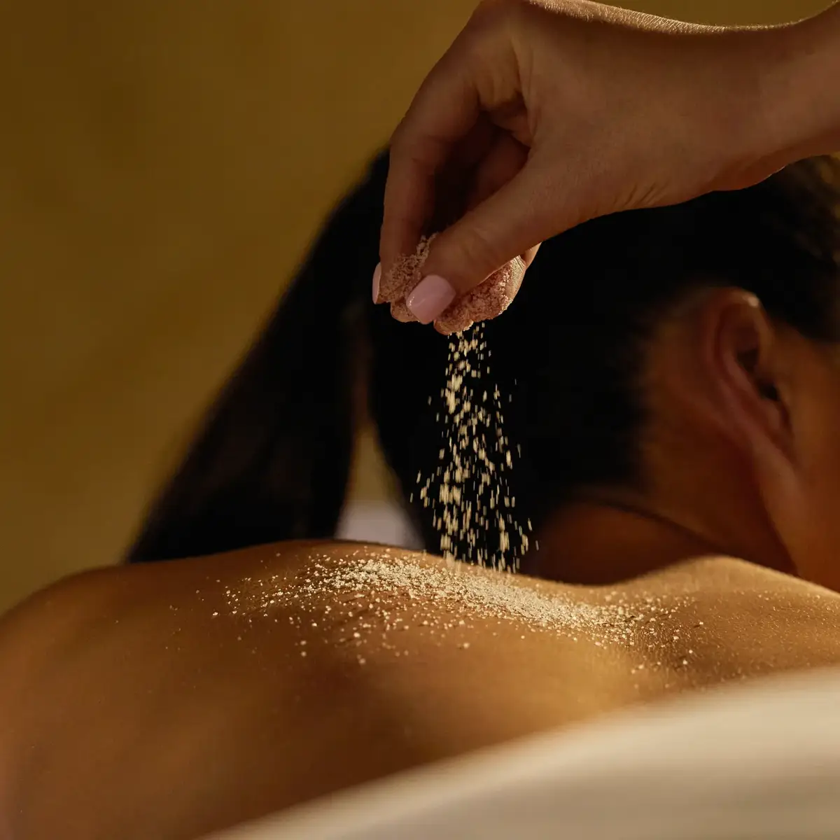 A hand sprinkles exfoliating salt onto a guest’s bare back during a spa treatment in a softly lit, relaxing setting.