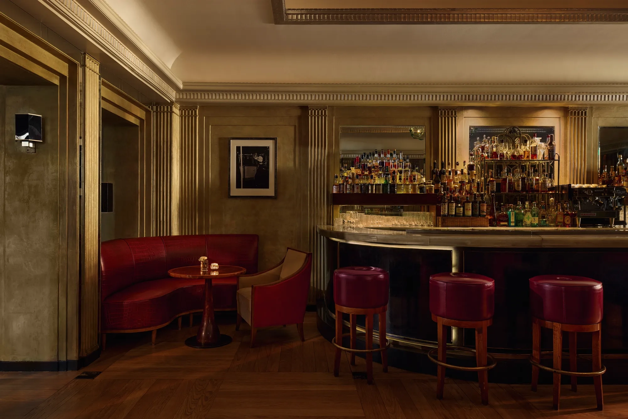 Elegant hotel bar with red leather seating, marble counter, and backlit spirits display.