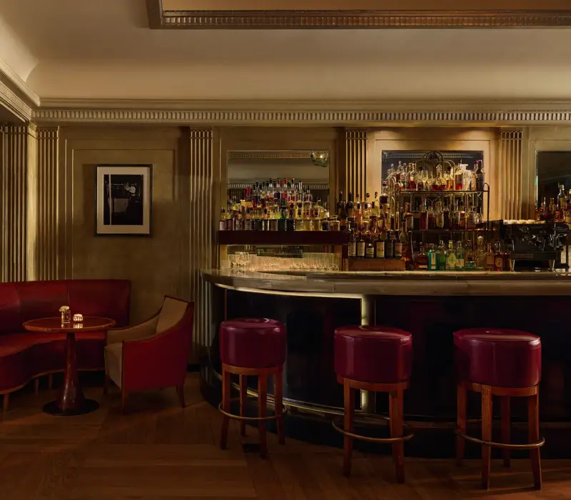 Elegant hotel bar with red leather seating, marble counter, and backlit spirits display.