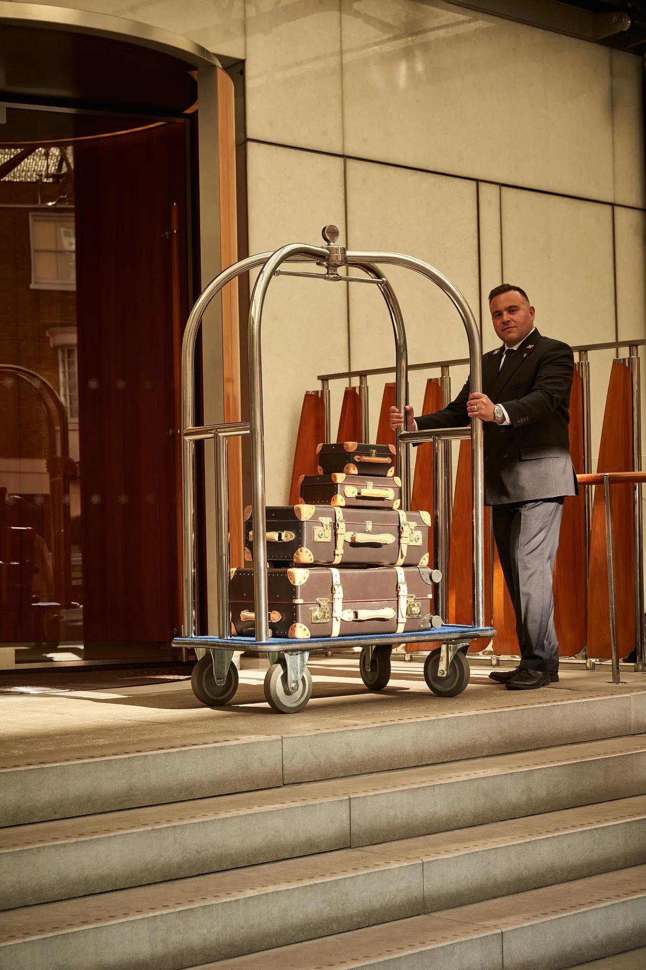 Doorman stands at a hotel entrance with a luggage trolley stacked with vintage-style suitcases.