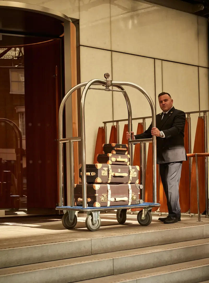 Doorman stands at a hotel entrance with a luggage trolley stacked with vintage-style suitcases.