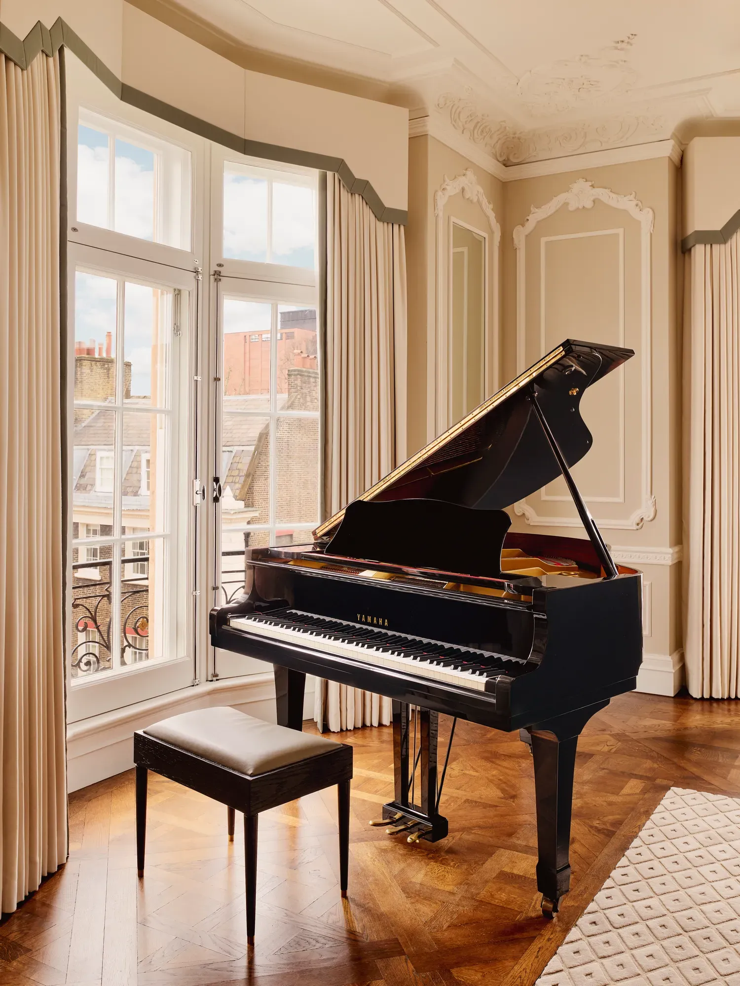 Elegant corner room with a black grand piano and stool set on parquet flooring, framed by tall windows with cream drapes and decorative wall panelling.