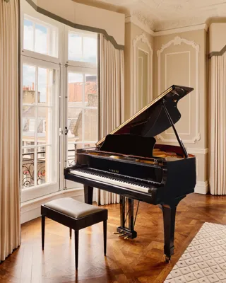 Elegant corner room with a black grand piano and stool set on parquet flooring, framed by tall windows with cream drapes and decorative wall panelling.