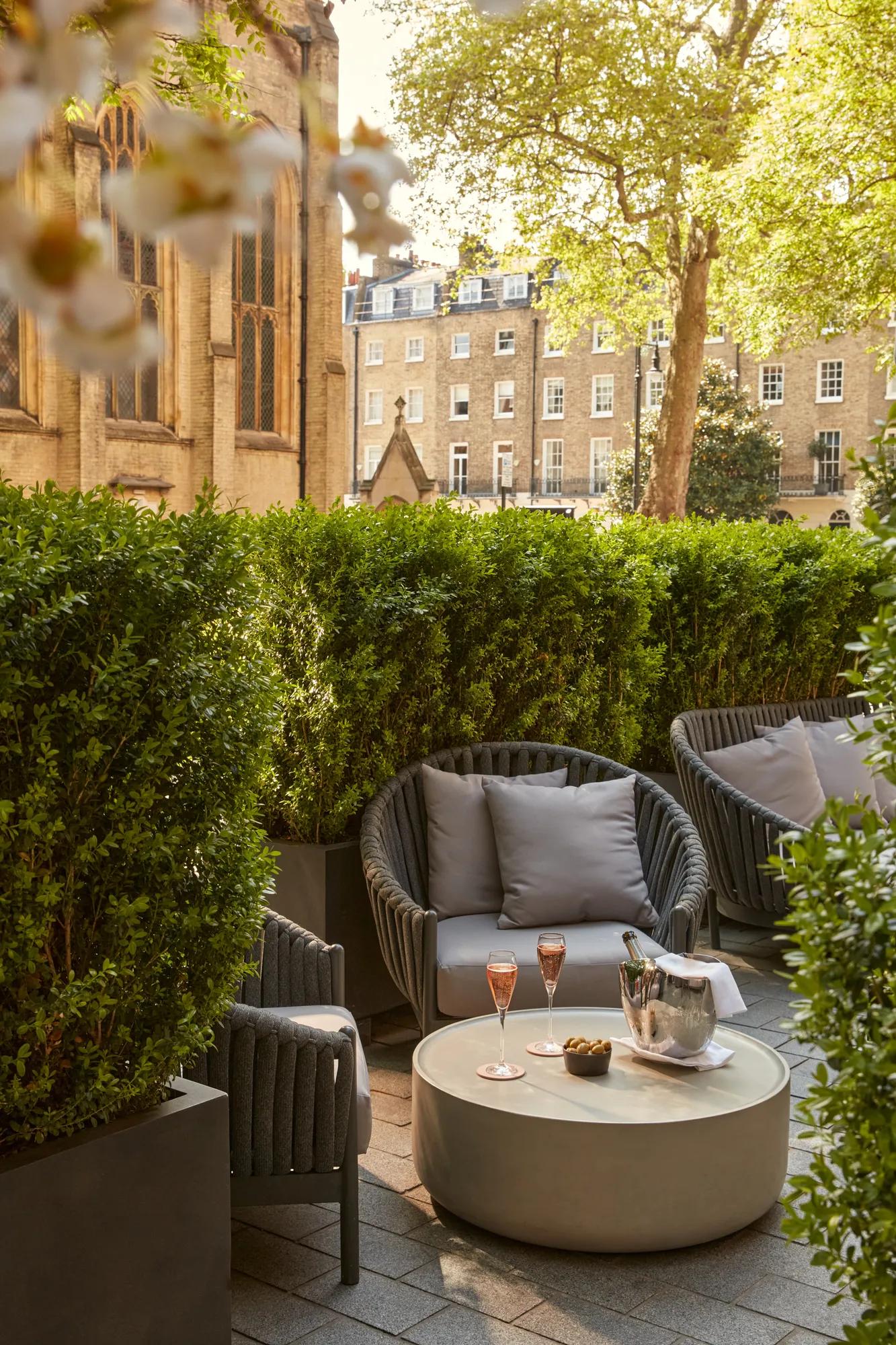 Outdoor seating area with grey cushioned chairs, low table with champagne, glasses, and snacks, beside historic church and greenery.