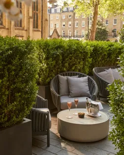 Outdoor seating area with grey cushioned chairs, low table with champagne, glasses, and snacks, beside historic church and greenery.