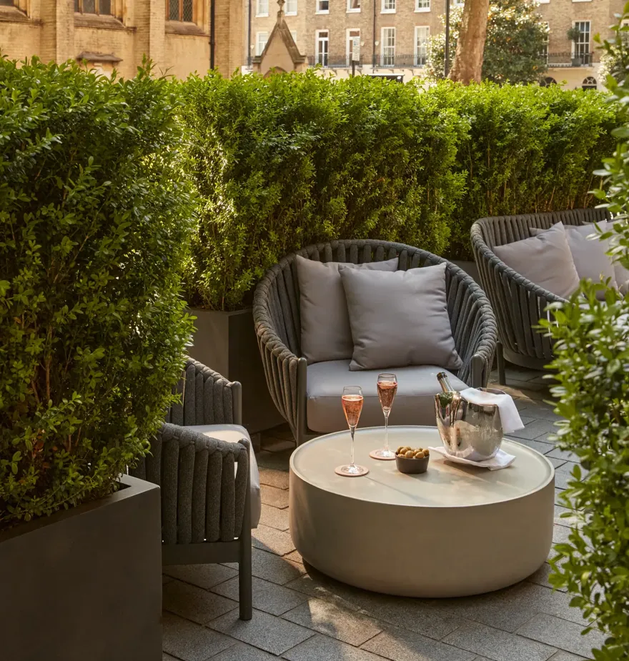 Outdoor seating area with grey cushioned chairs, low table with champagne, glasses, and snacks, beside historic church and greenery.
