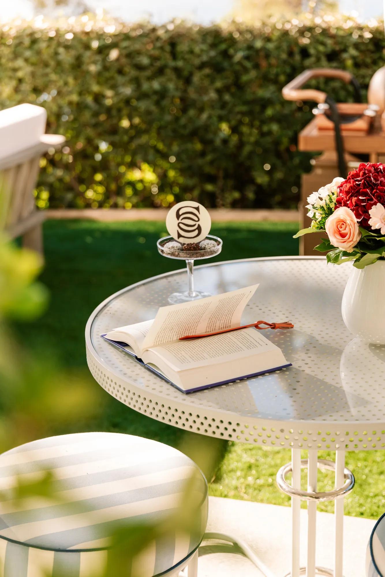 Open book on round outdoor table with glass dessert and flowers, set in sunny garden with lawn and greenery in background.