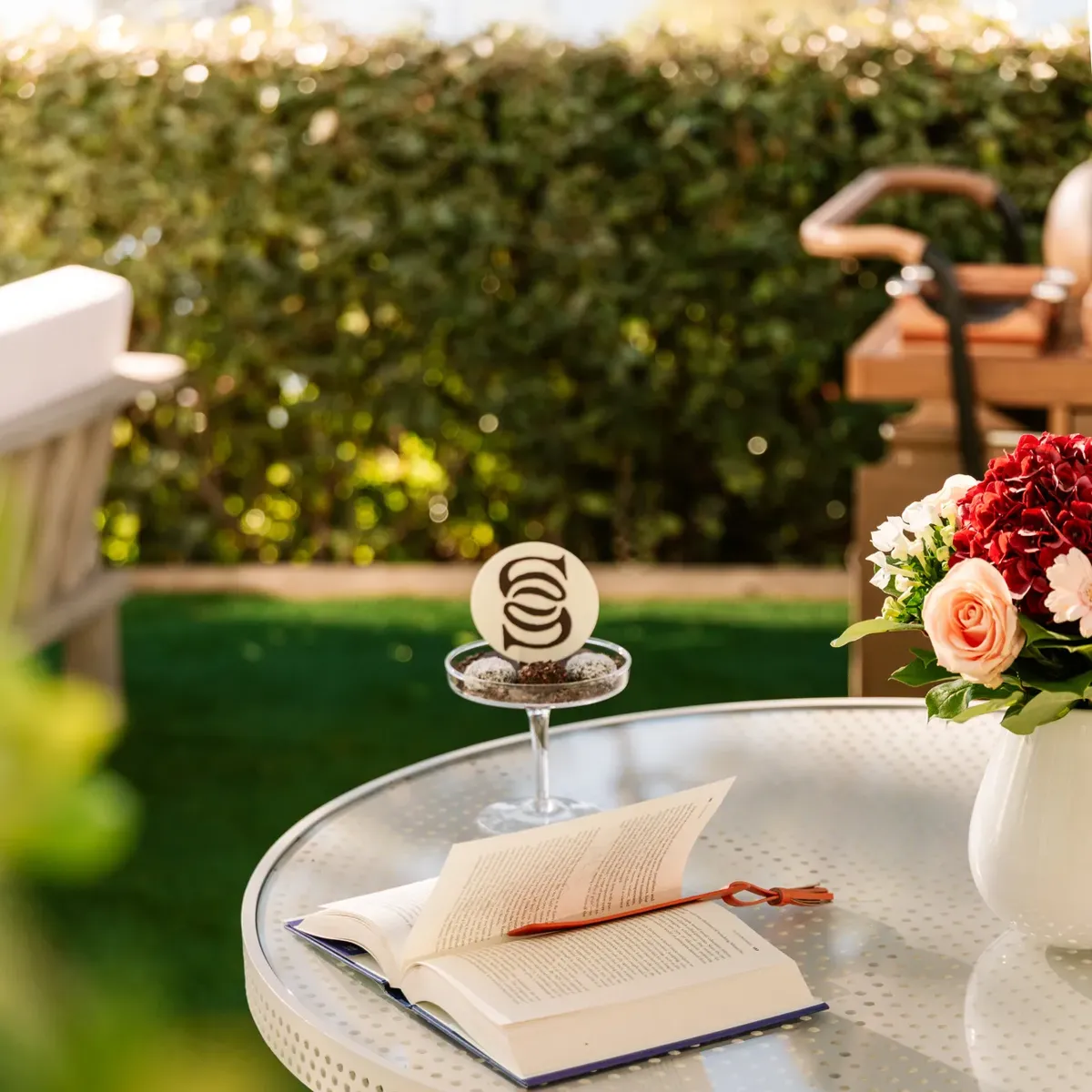 Open book on round outdoor table with glass dessert and flowers, set in sunny garden with lawn and greenery in background.