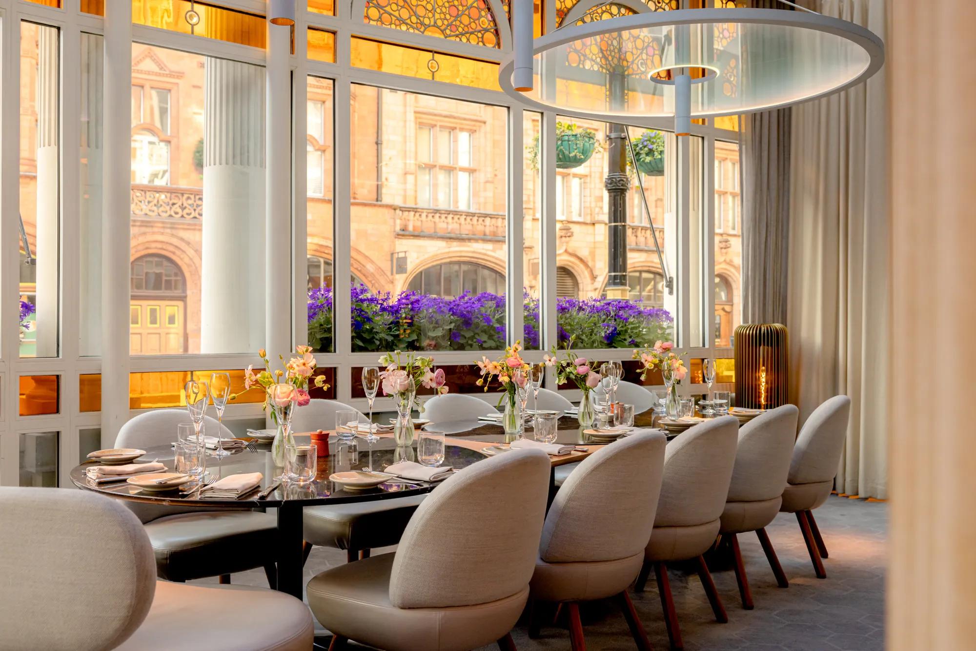 Elegant restaurant dining area with a long set table, upholstered chairs, and large windows overlooking a historic building.