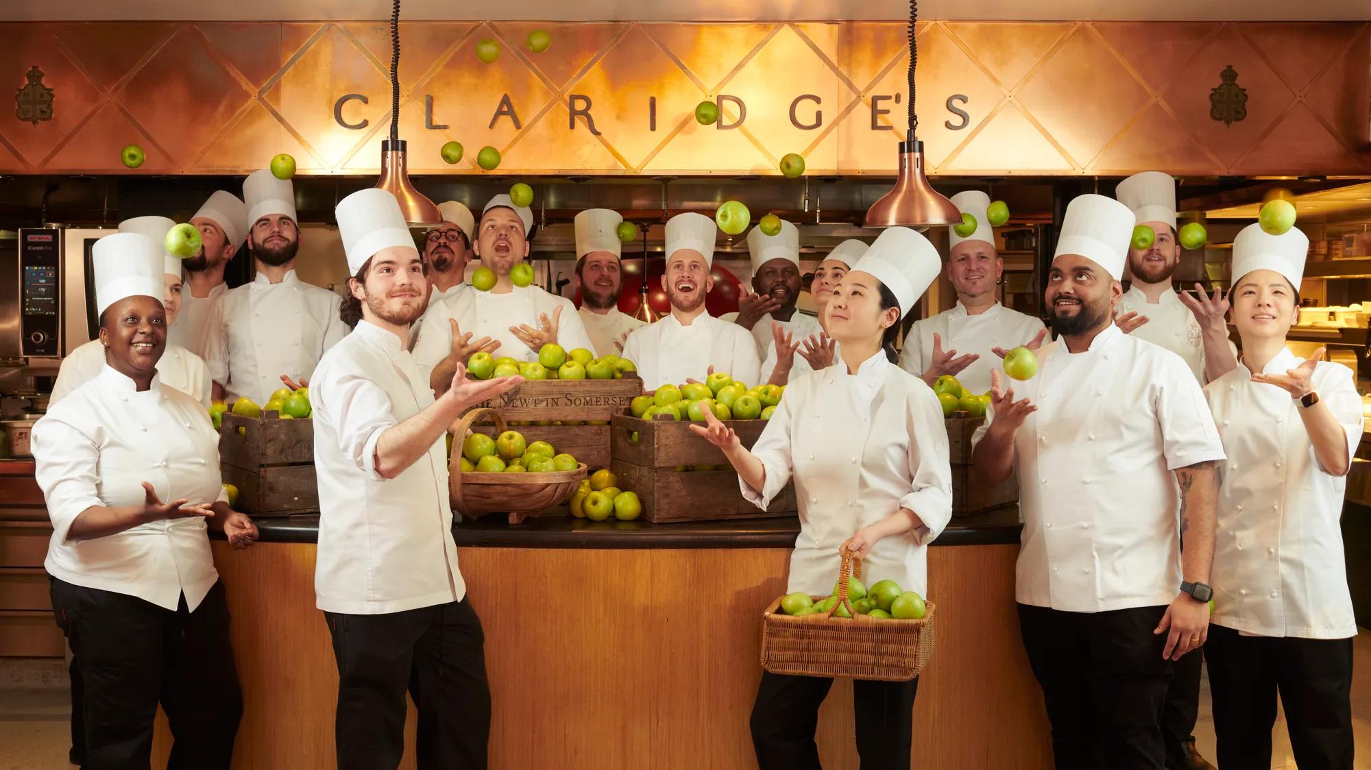 Chefs in white uniforms tossing green apples in kitchen beneath Claridge's sign.