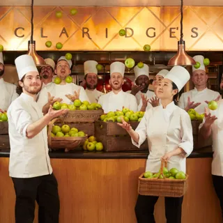 Chefs in white uniforms tossing green apples in kitchen beneath Claridge's sign.