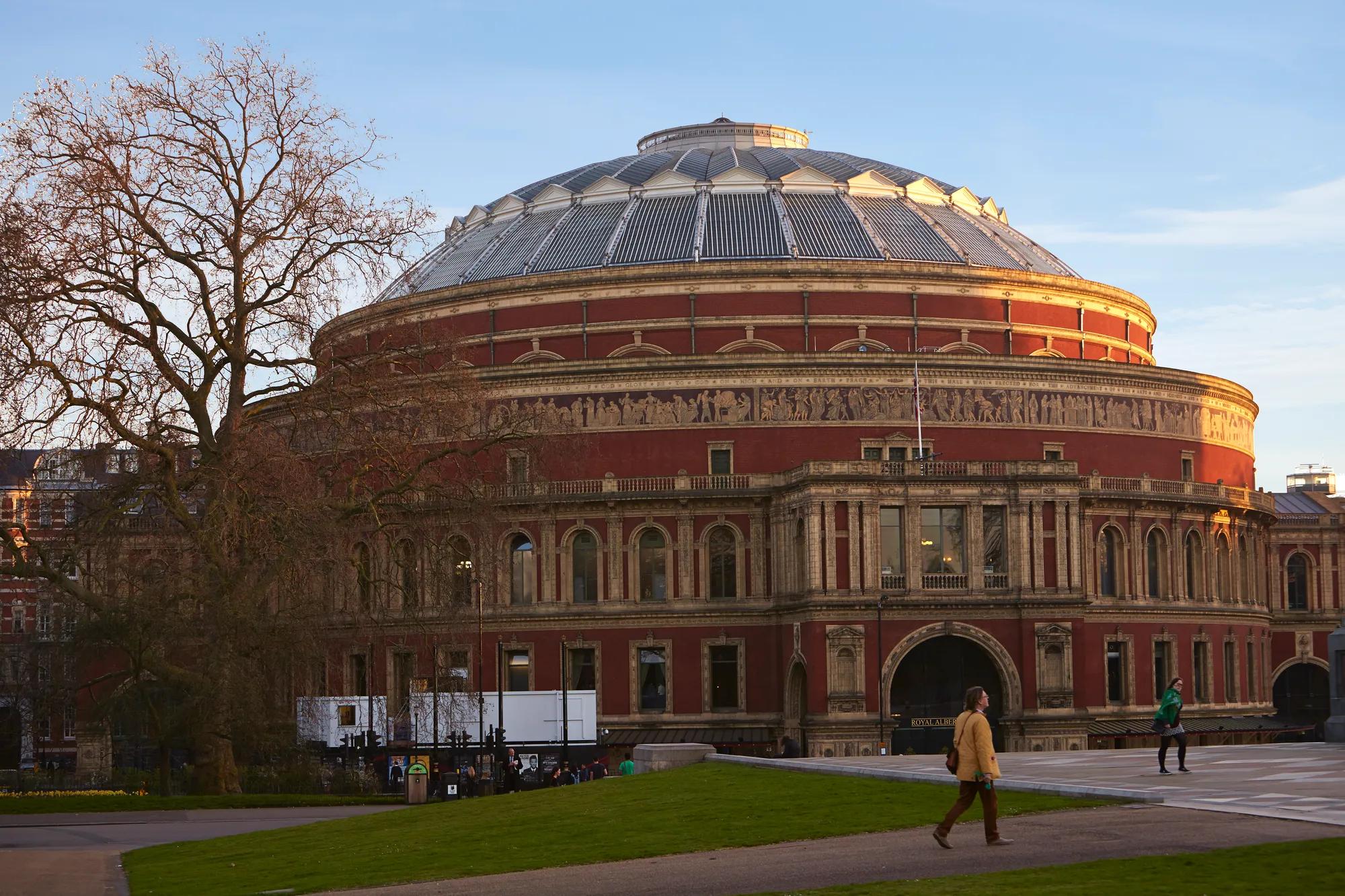 Exterior of the Royal Albert Hall, a circular red-brick building with domed roof, viewed from a park with pedestrians nearby.