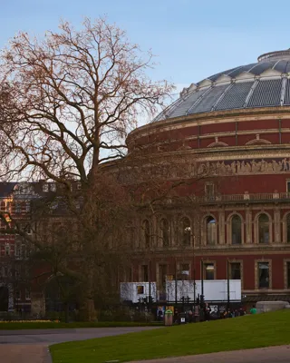 Exterior of the Royal Albert Hall, a circular red-brick building with domed roof, viewed from a park with pedestrians nearby.