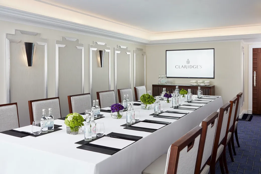 Modern meeting room at Claridge’s arranged with a long conference table, elegant chairs, bottled water, notepads, and a screen displaying the hotel’s logo.
