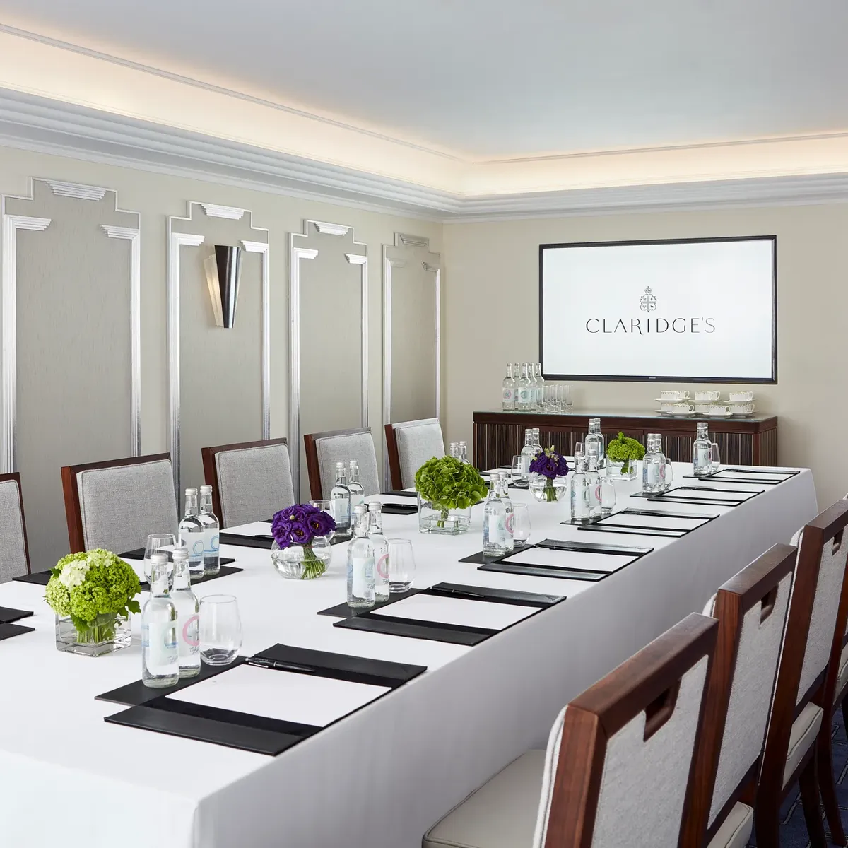 Modern meeting room at Claridge’s arranged with a long conference table, elegant chairs, bottled water, notepads, and a screen displaying the hotel’s logo.