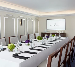 Modern meeting room at Claridge’s arranged with a long conference table, elegant chairs, bottled water, notepads, and a screen displaying the hotel’s logo.