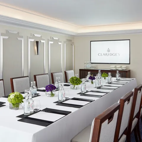 Modern meeting room at Claridge’s arranged with a long conference table, elegant chairs, bottled water, notepads, and a screen displaying the hotel’s logo.